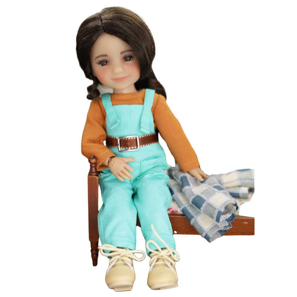Doll wearing a brown sweater and turquoise overalls sitting on a chair with a plaid blanket.